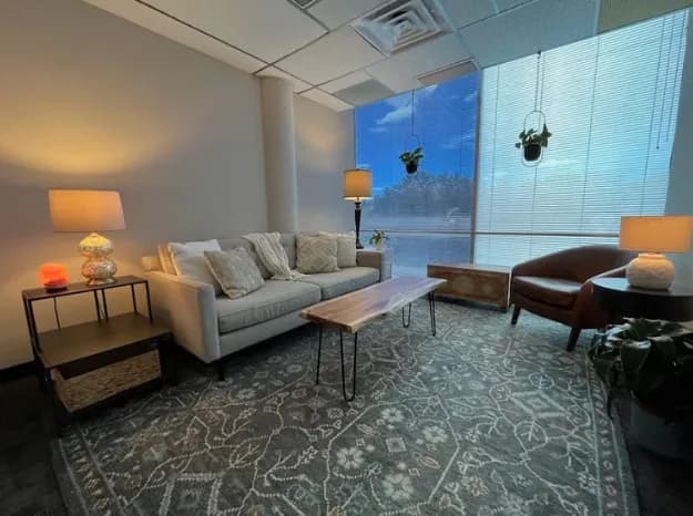 Therapy room with a sofa, armchair, and hanging plants beside a large window